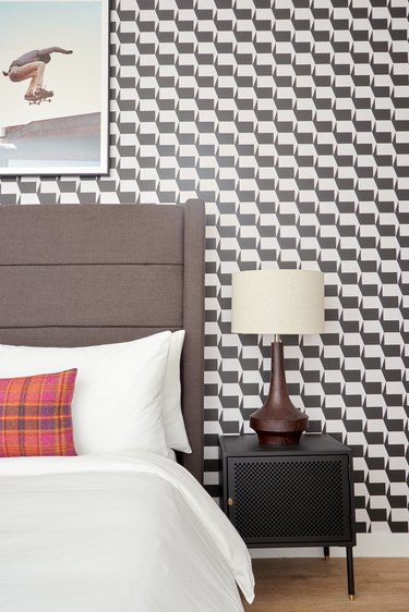 Eclectic bedroom with dark brown fabric headboard, black and white pattern wallpaper, lamp, nightstand, plaid red pillow.